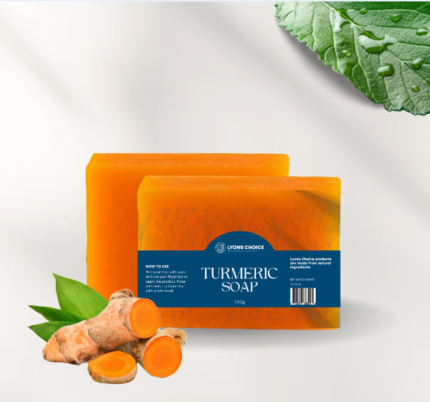This is an image of turmeric sopa - a product listed on page Wholesale - Lyons Choice woocommerce store. The image features a packaging of soap with turmeric yellow color of cuboidal packing and the name of the product written on blue color background and two pieces of fresh turmeric the green leaves also featuring on the left side of the product.